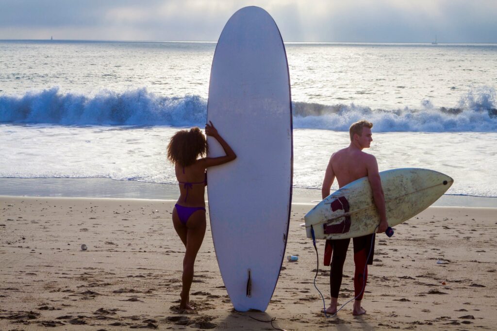 Woman next to a tall longboard and a man holding a small shortboard on the beach.