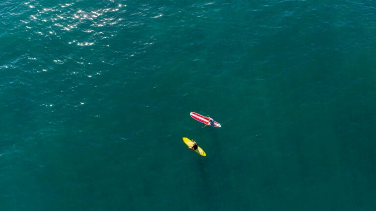 2 surfers floating in the ocean. 1 surfer is on a hard top surfboard while the other is on a soft top surfboard